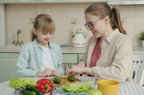 Young woman is cooking salad and feeding cute little daughter with cucumber slices in kitchen, family is talking and having good time together.