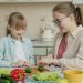 Young woman is cooking salad and feeding cute little daughter with cucumber slices in kitchen, family is talking and having good time together.